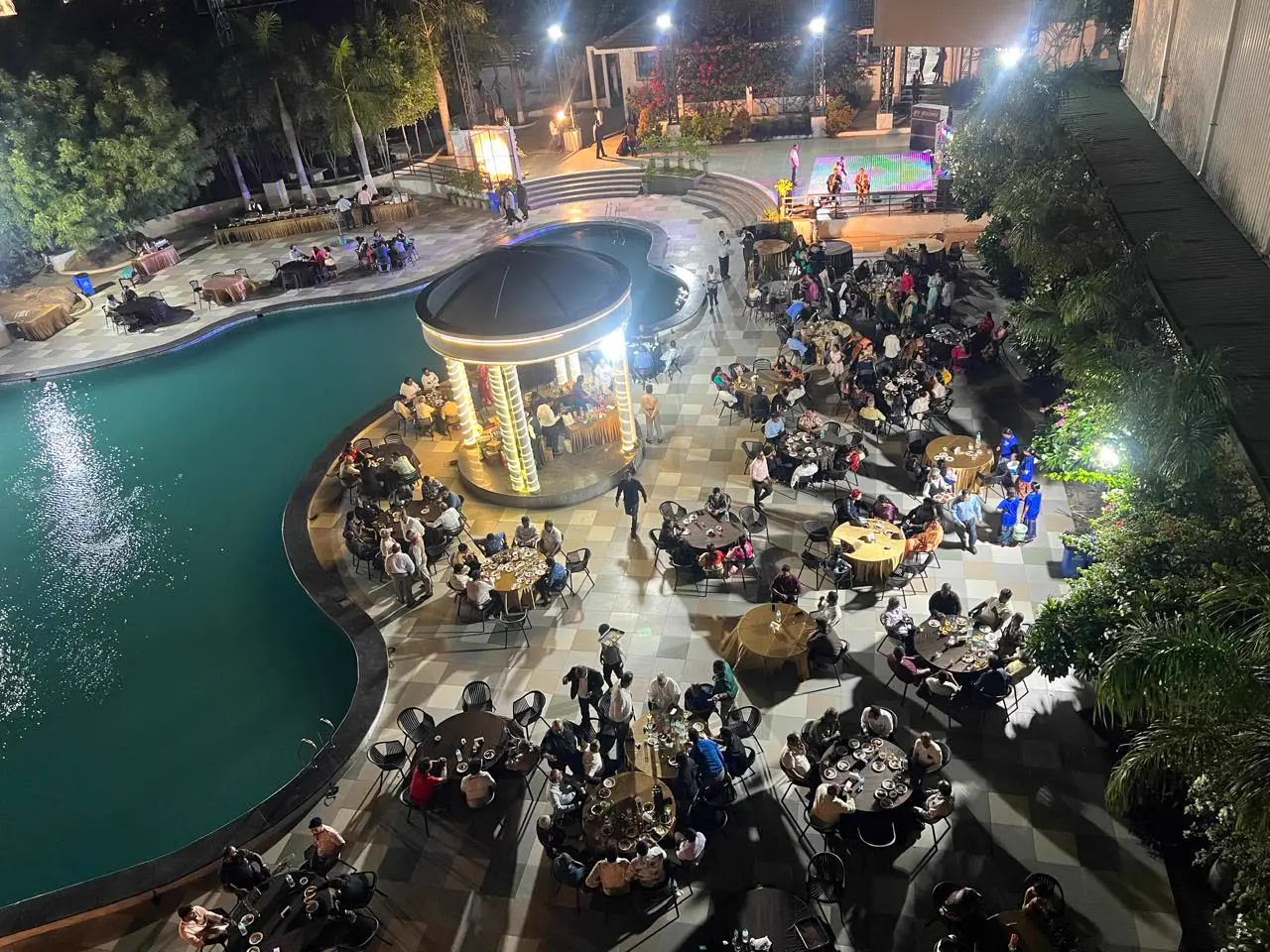 A banquet at night around the lit pool and gazebo, with a stage beyond