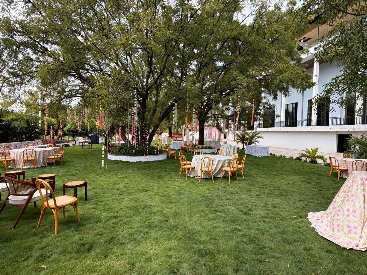 A lawn under big trees set with chairs and tables, floral strands hanging from the branches